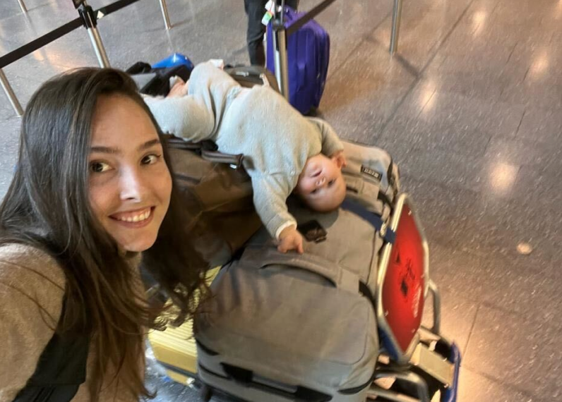 Un compañero sonriente junto a una niña tumbada bocabajo sobre maletas en el aeropuerto.